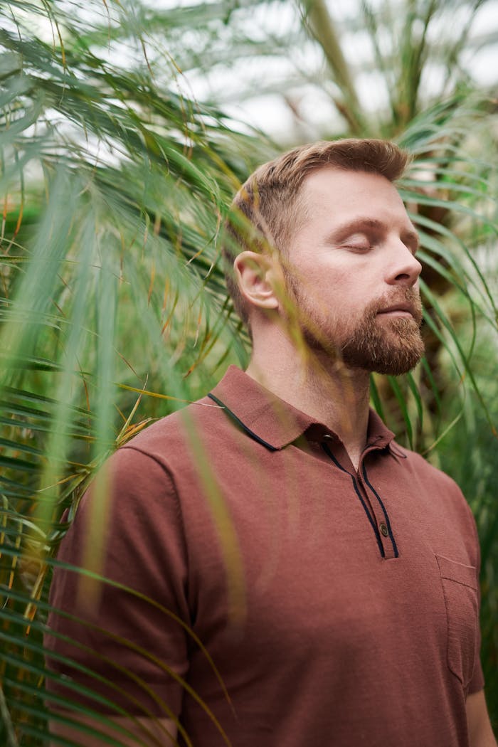 A man in a polo shirt enjoys a peaceful moment with closed eyes among green leaves.