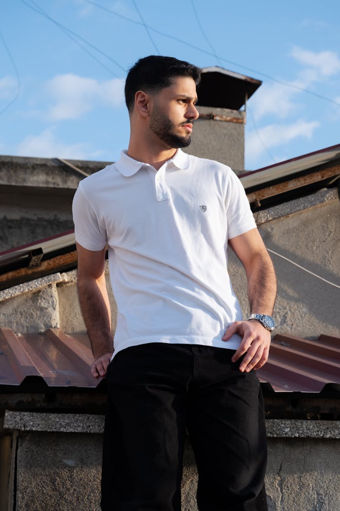 Young man in casual attire posing confidently on a rooftop with a clear blue sky backdrop.